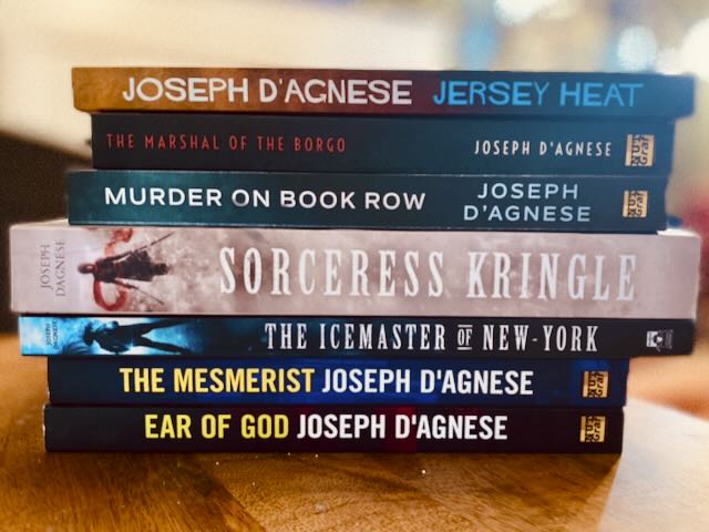 Joe's Paperback Writer Bundle book cover by Joseph D'Agnese, featuring a real-life photo of seven paperback books stacked in a pile, including titles like Jersey Heat, The Marshal of the Borgo, and Sorceress Kringle. The image represents a massive multi-genre collection of 2,200 pages of thriller, mystery, and fantasy novels.