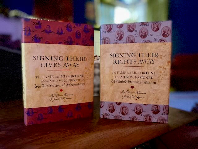 Signing Their Lives Away and Signing Their Rights Away hardcover books standing upright on a desk, displaying their dust jackets with thumbnail portraits of the signers. Two autographed editions by Joseph D'Agnese and Denise Kiernan.